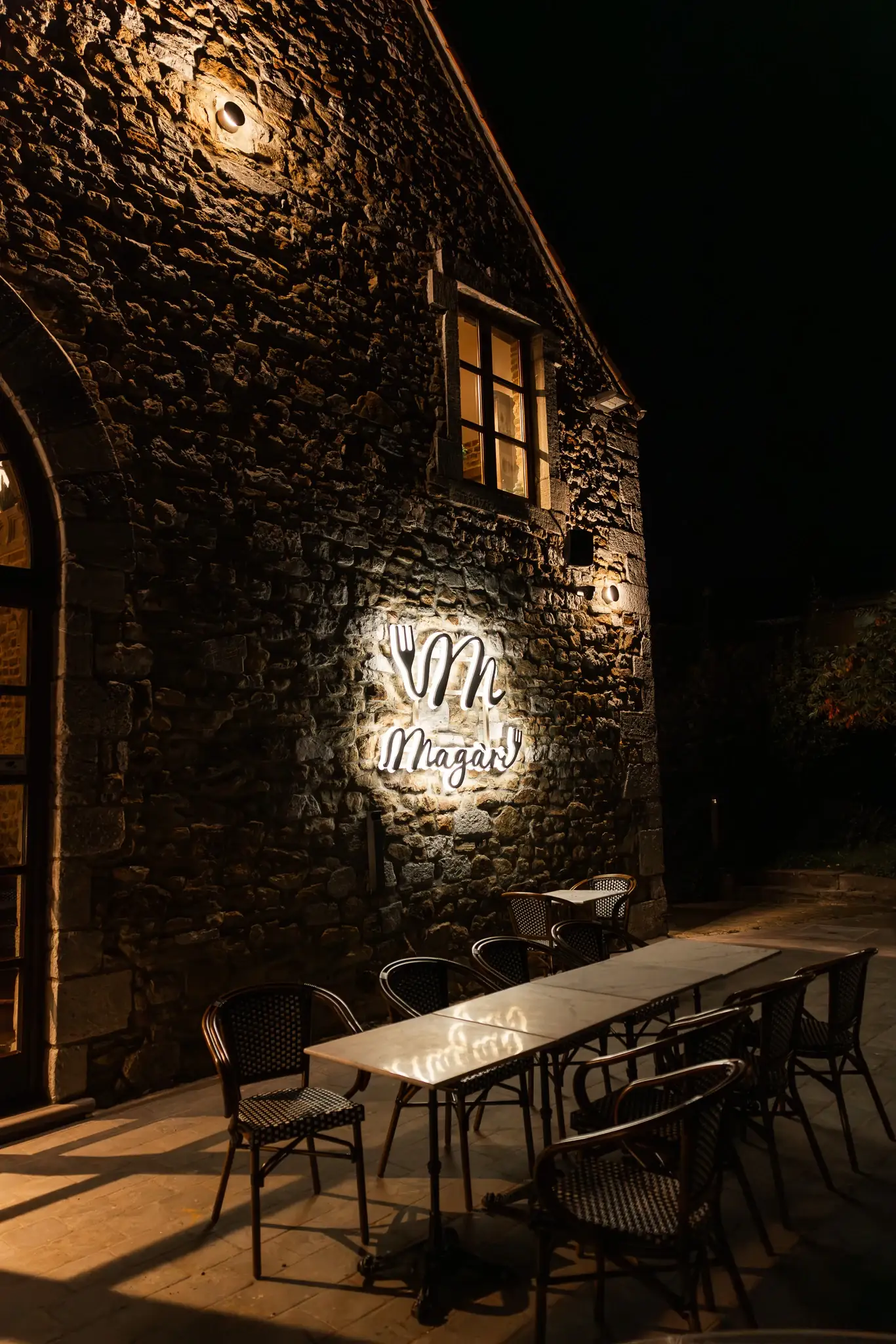 La façade en pierre et l’enseigne lumineuse du restaurant Magari, de nuit